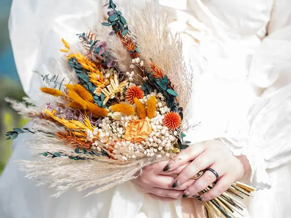 Close-up of hands holding a boho-style dried flower bouquet with pampas grass, eucalyptus, and orange accents, set against a white long-sleeved dress.