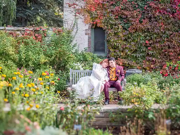 A bride in a white dress resting her head on her partner's shoulder while sitting on a wooden bench surrounded by colorful flowers and red foliage.