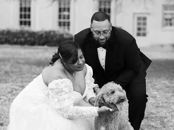 Black and white photograph of a bride and groom smiling while petting their dog outdoors