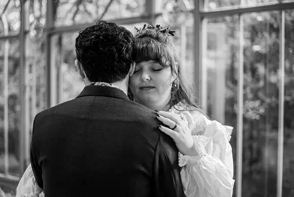 Black and white photograph of a bride with her eyes closed resting her hand on her groom's shoulder inside a glass greenhouse
