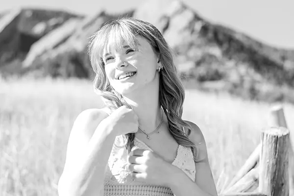 Black and white outdoor portrait of a smiling young woman with bangs standing in a field near a wooden fence with mountains behind her